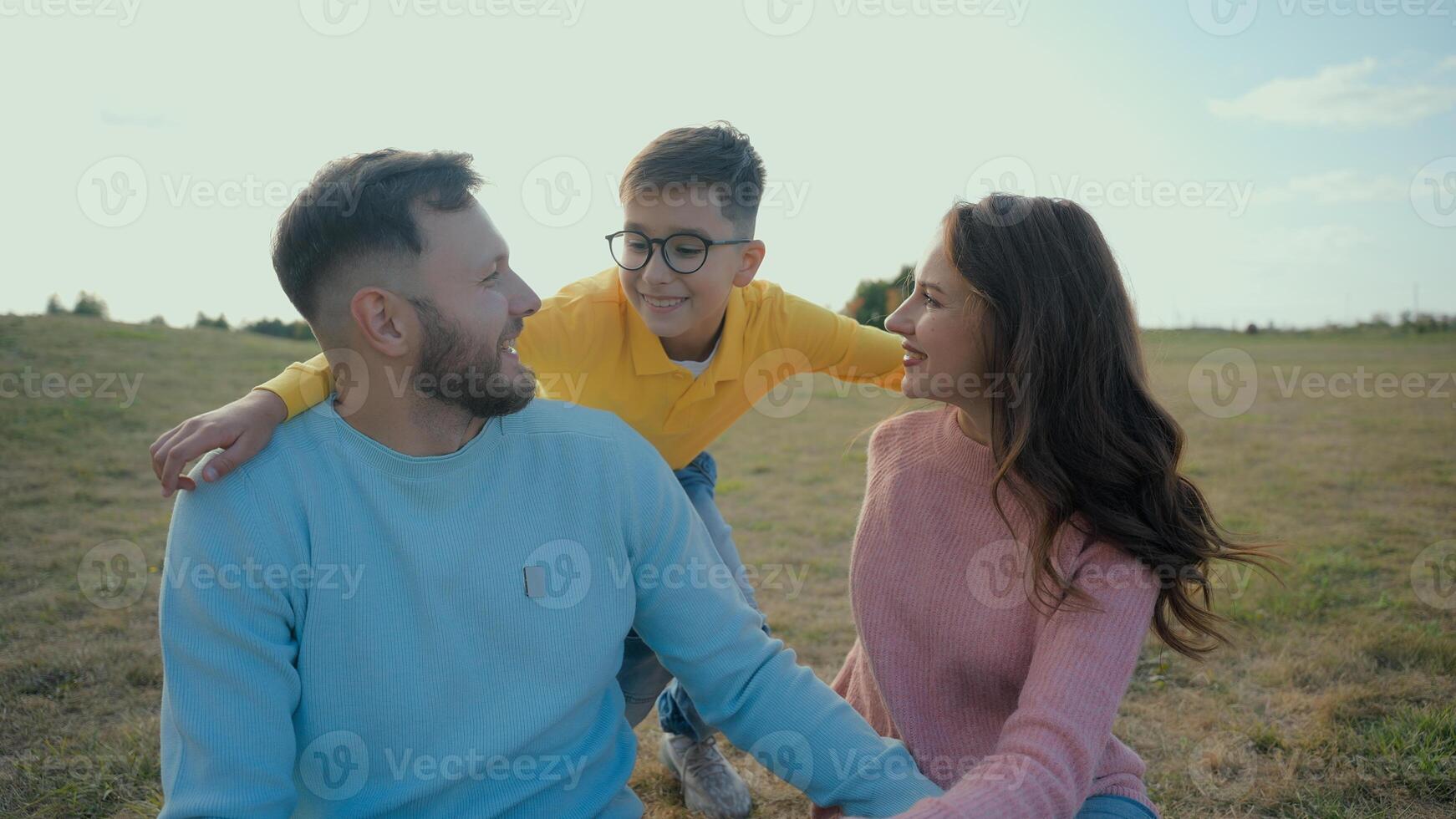 Joyful Caucasian family picnic married couple mother female woman father dad guy man male hug happy little boy child kid son walking to parents embrace cuddle together looking at camera nature meadow photo