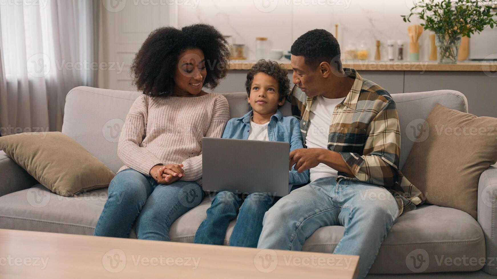 Happy married woman female mom male dad man African American family couple talking explain notebook pc software use internet app little boy child son kid living room couch. Parental control technology photo