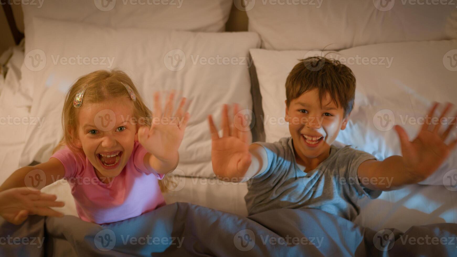 Two happy smiling funny friendly Caucasian kids little son brother boy daughter sister girl children lying bed pillows surprising waving hands greeting hello gesture blankets evening bedroom indoors photo