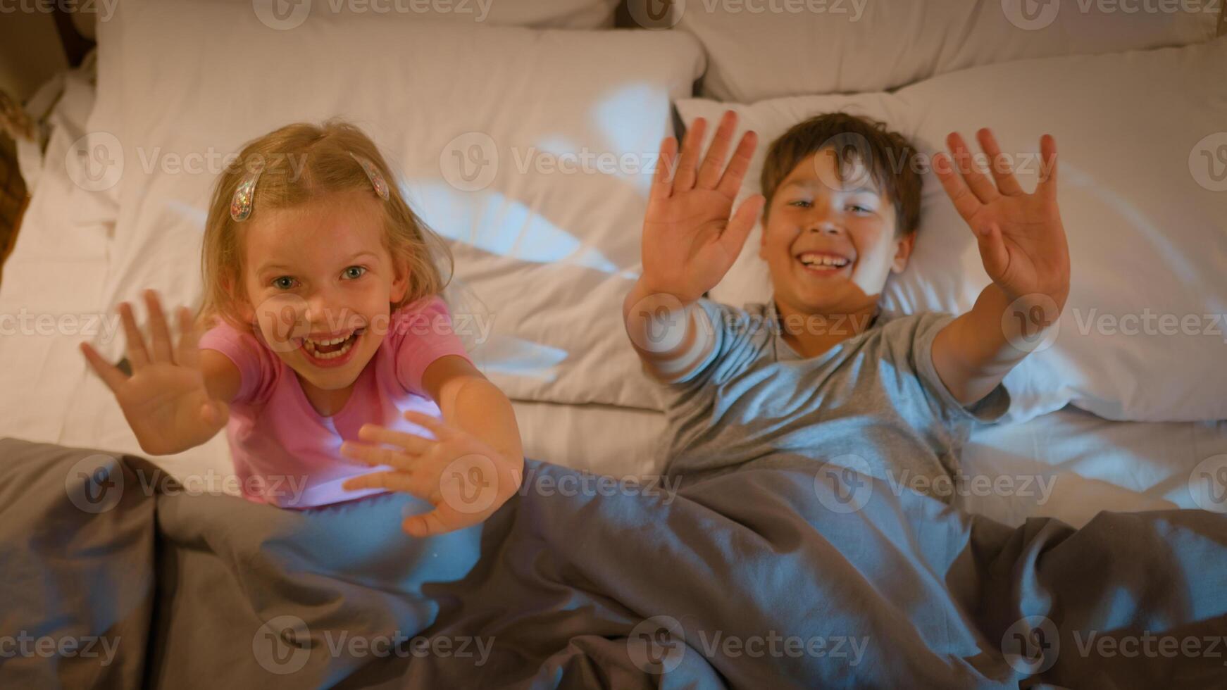 Two happy smiling funny friendly Caucasian kids little son brother boy daughter sister girl children lying bed pillows surprising waving hands greeting hello gesture blankets evening bedroom indoors photo