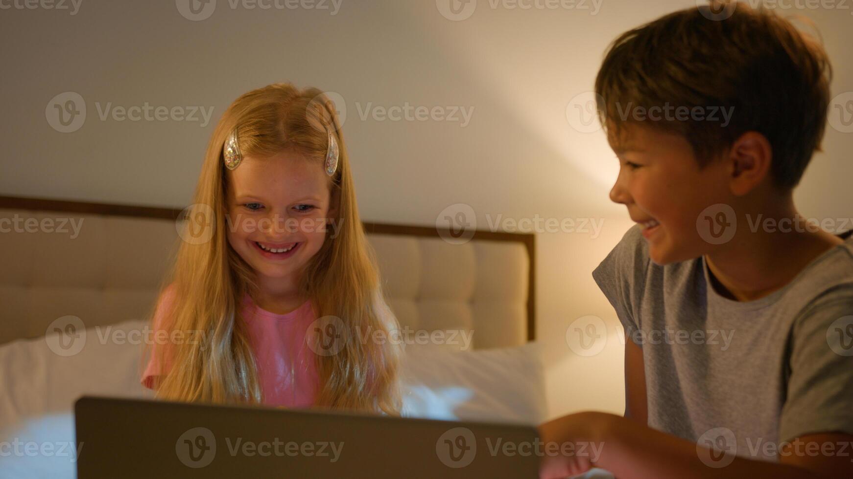 Happy little Caucasian children kids siblings family laugh kids watching online use laptop computer boy son girl daughter friends cartoon movie together bed night insomnia awake inside bedroom photo