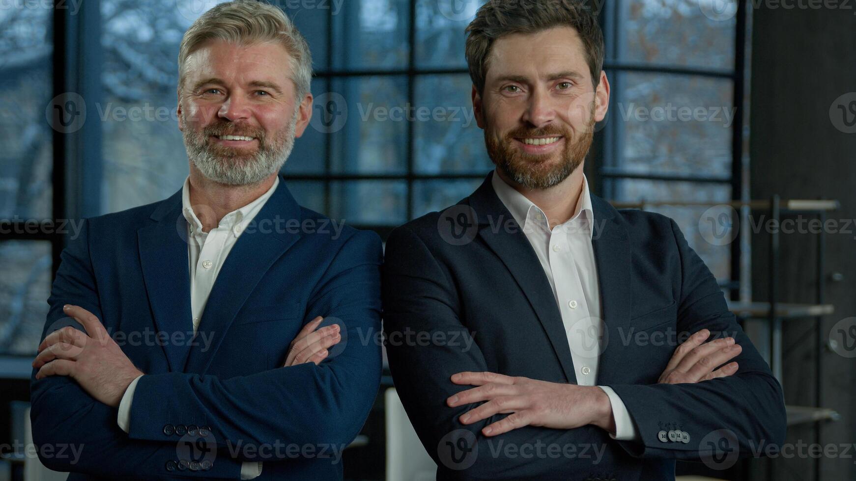 Two successful smiling businessmen posing together with crossed arms young and mature man business partners colleagues corporate employees professional managers standing in office looking at camera photo
