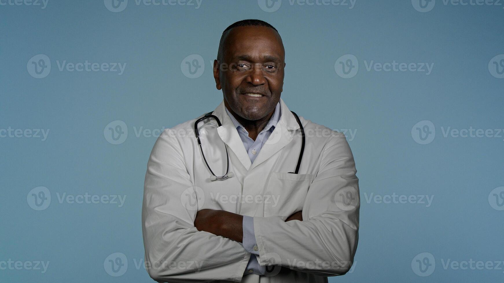 Mature confident happy doctor standing in gray background studio with arms crossed glad smiling african american man professional therapist physician in medical gown posing indoors looking at camera photo