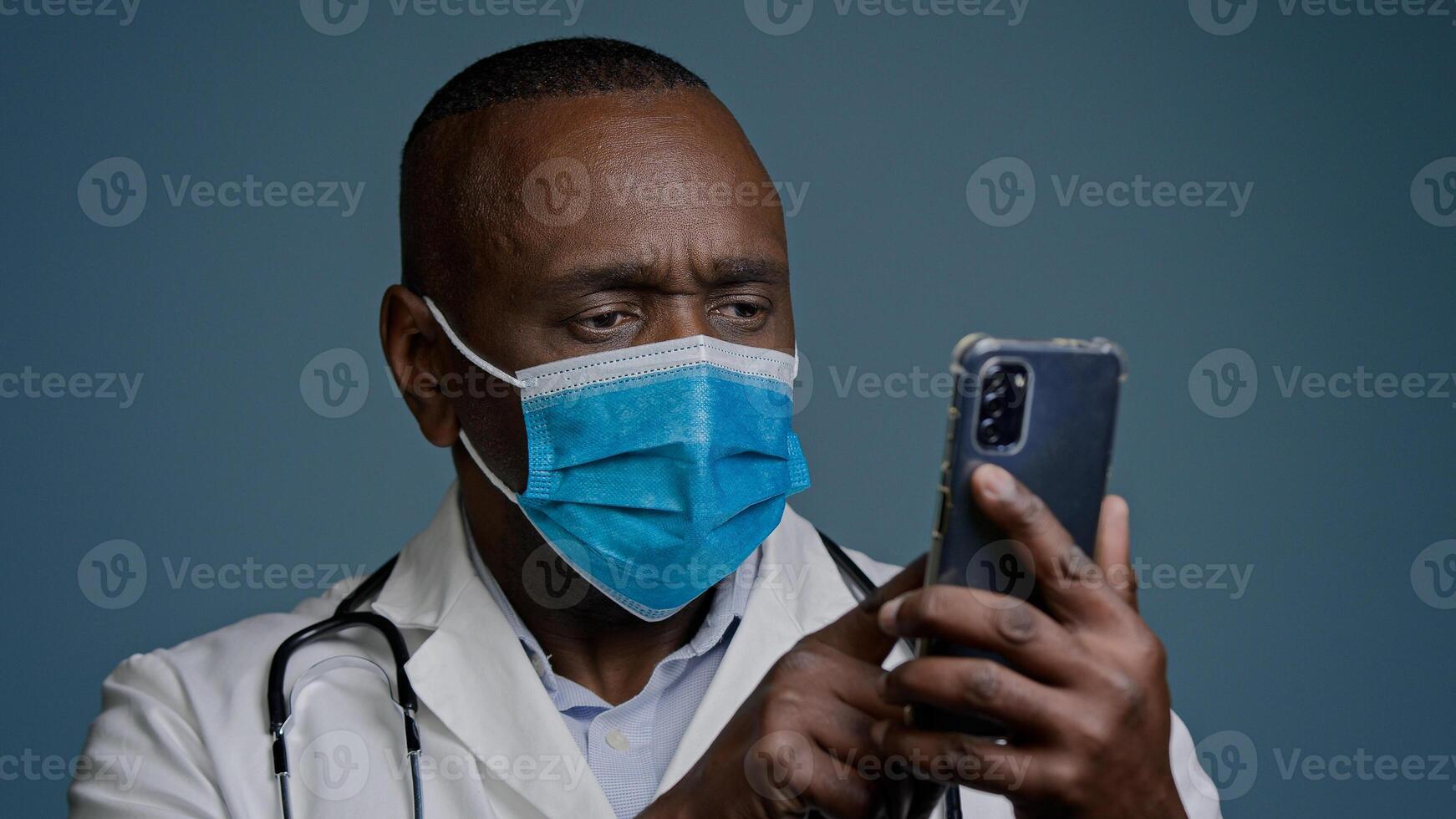 Close-up experienced mature doctor in protective mask and white medical gown reads message on mobile phone checks information on Internet concentrated man points to smartphone screen nods approval photo