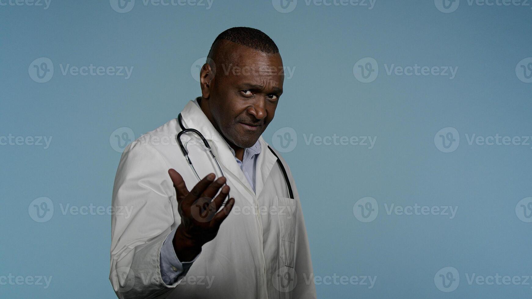 African american man in medical gown walking in gray background studio male doctor hospital employee invites patients to use professional service medicare waving hand makes come here beckoning gesture photo