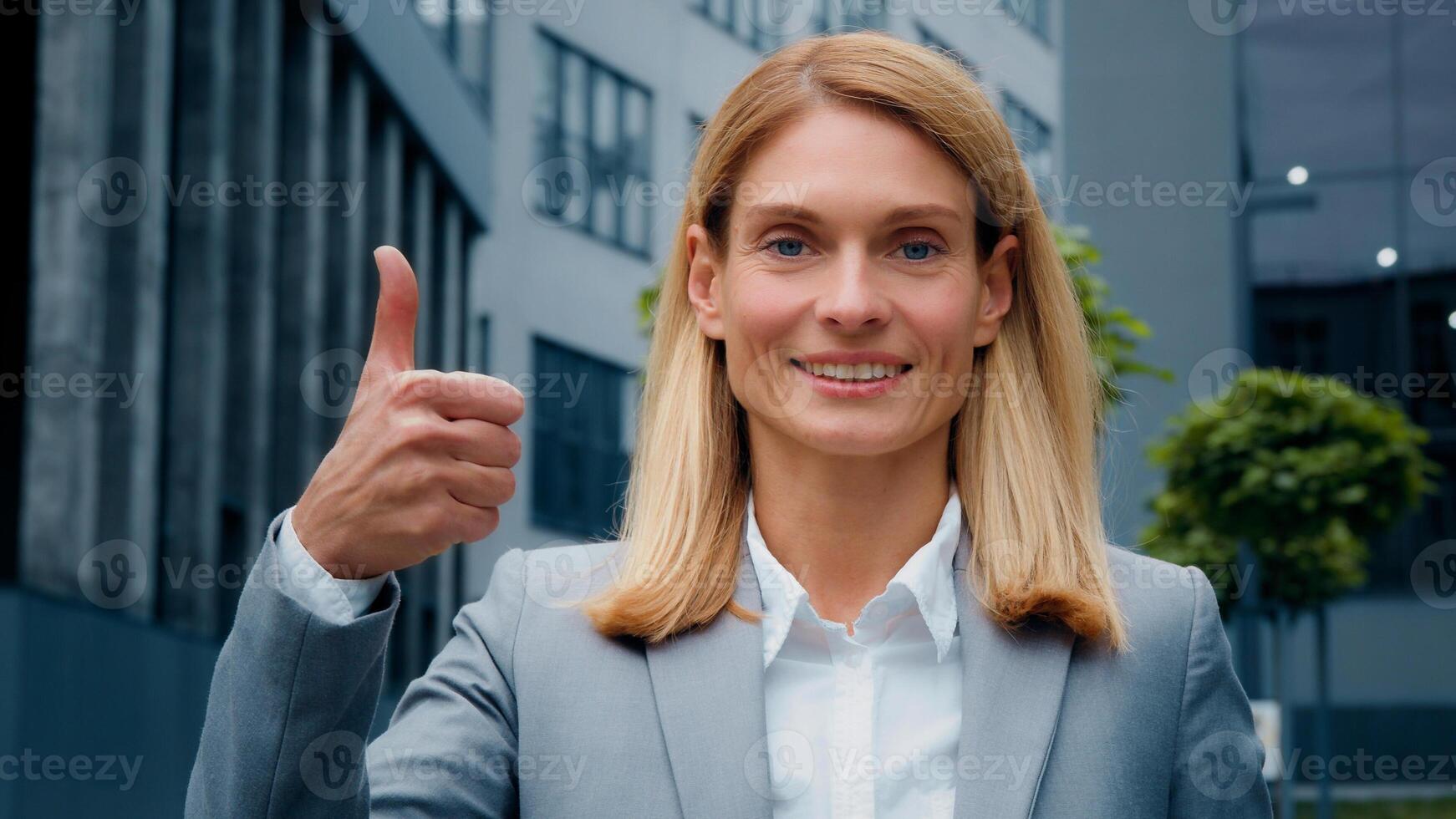 Close-up young smiling businesswoman in suit standing outdoors against backdrop office building looking at camera showing thumb up gesture approval sign achievement agreement symbol shows good result photo