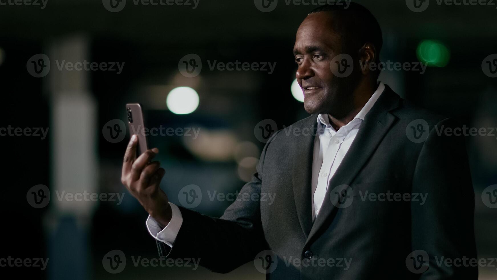 Mature african american businessman talks via link using mobile phone standing in dark room puzzled man communicates on online conference answering business call looks at cellphone screen photo