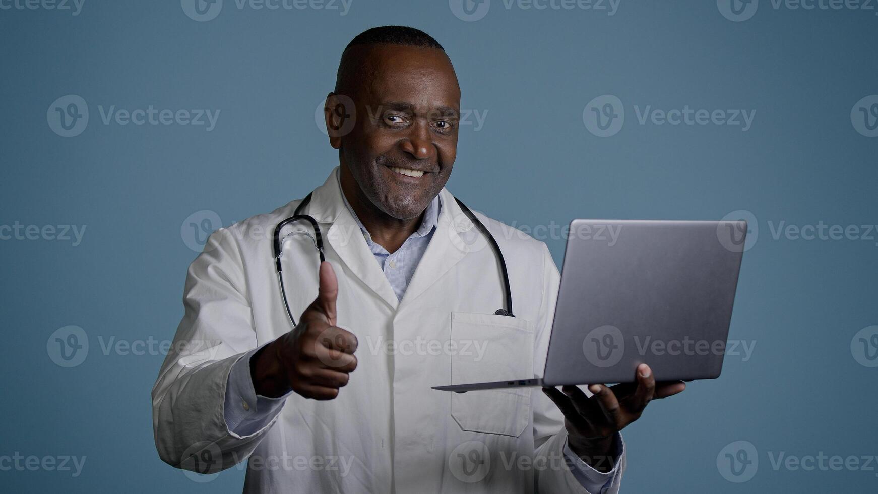 Mature man stands in studio on gray background professional doctor in medical gown checks tests using laptop consults patient online showing thumb up gesture approval good result healthcare concept photo