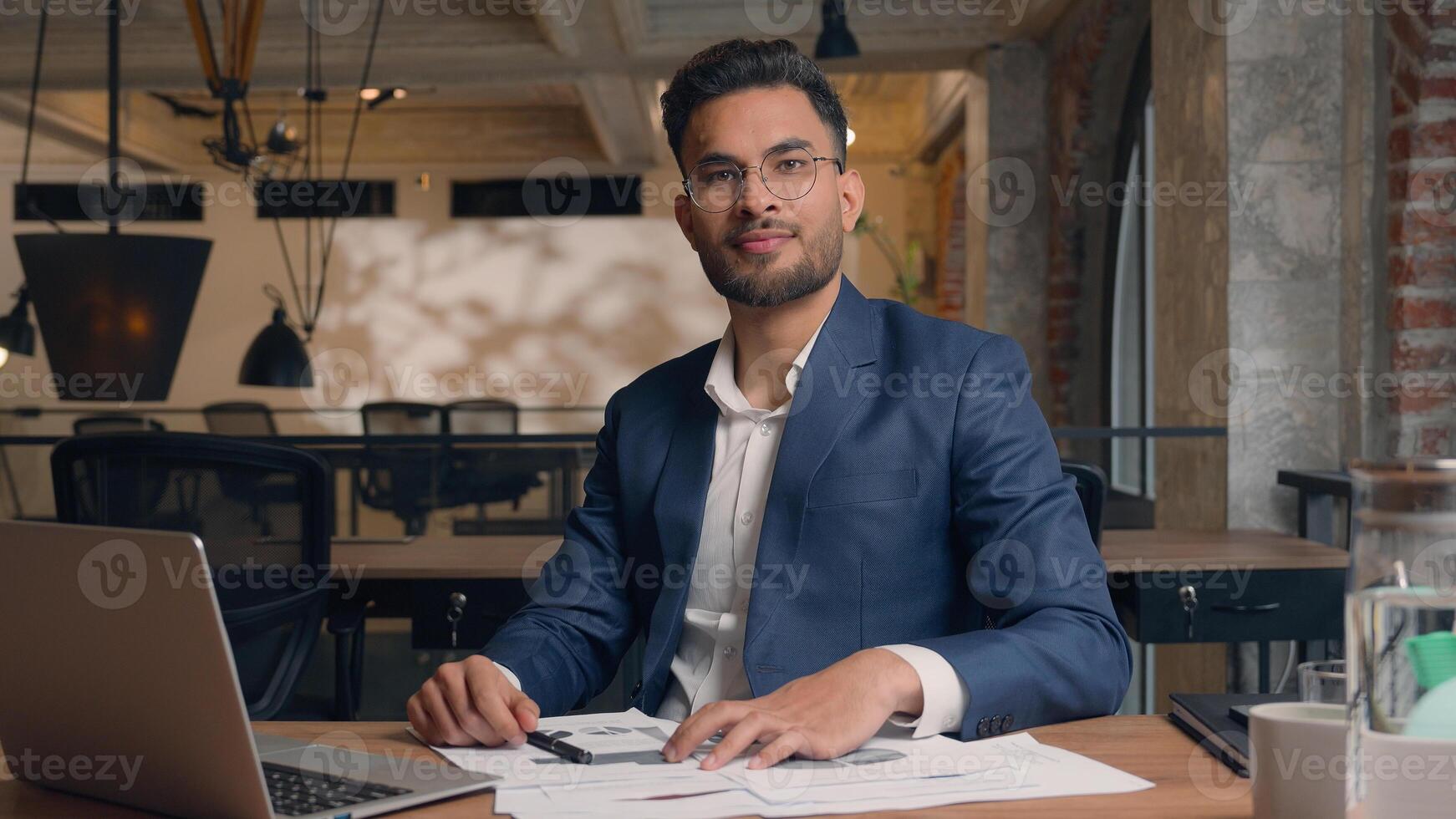 Indian businessman male entrepreneur leader corporate manager work computer laptop write documents paperwork on desk Arabian guy business man happy positive smile guy looking at camera in office cafe photo