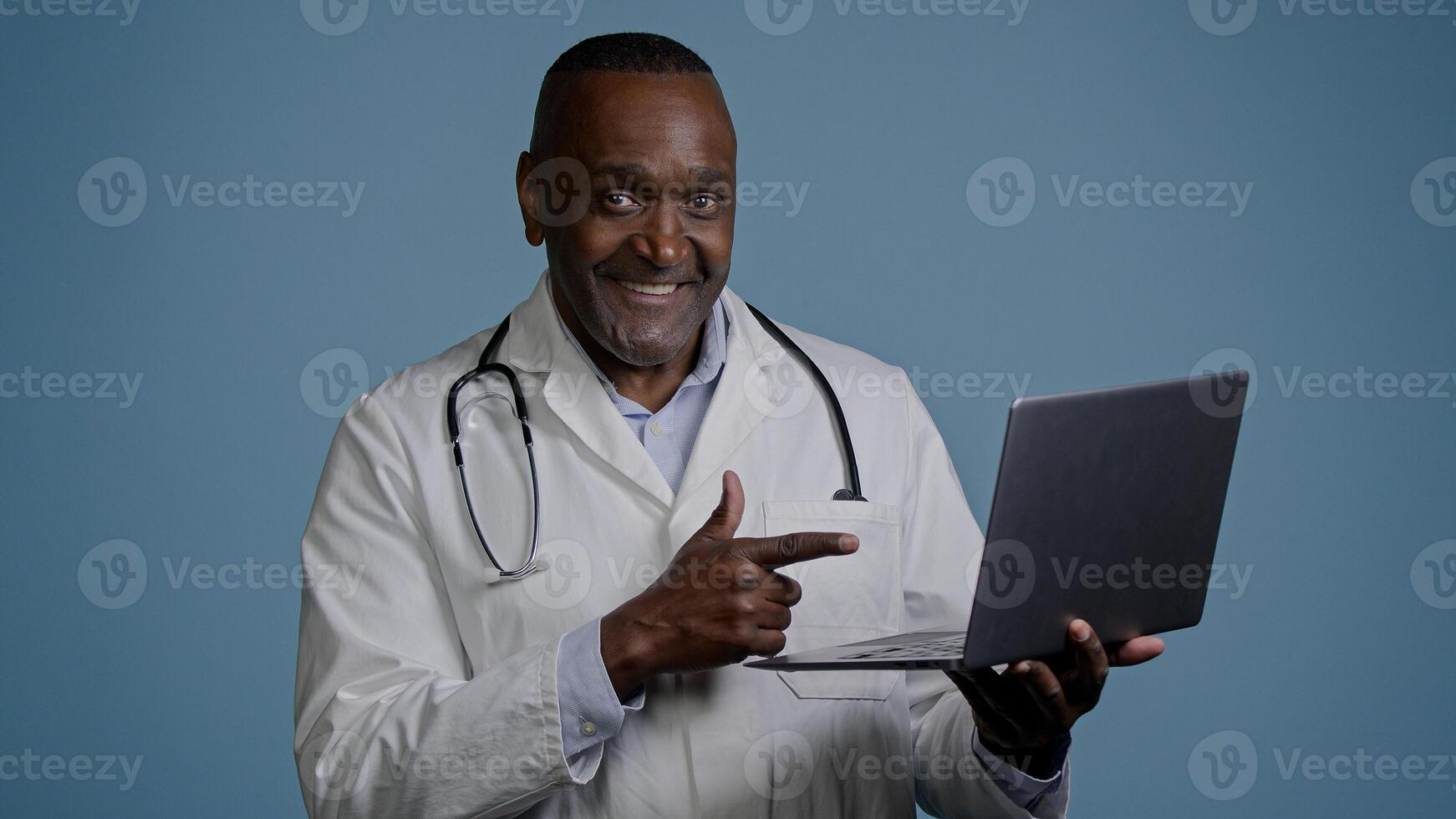 African american man doctor therapist ethnic male surgeon in white coat in blue background studio working on laptop consulting patient online remote diagnostics take useful information from internet photo