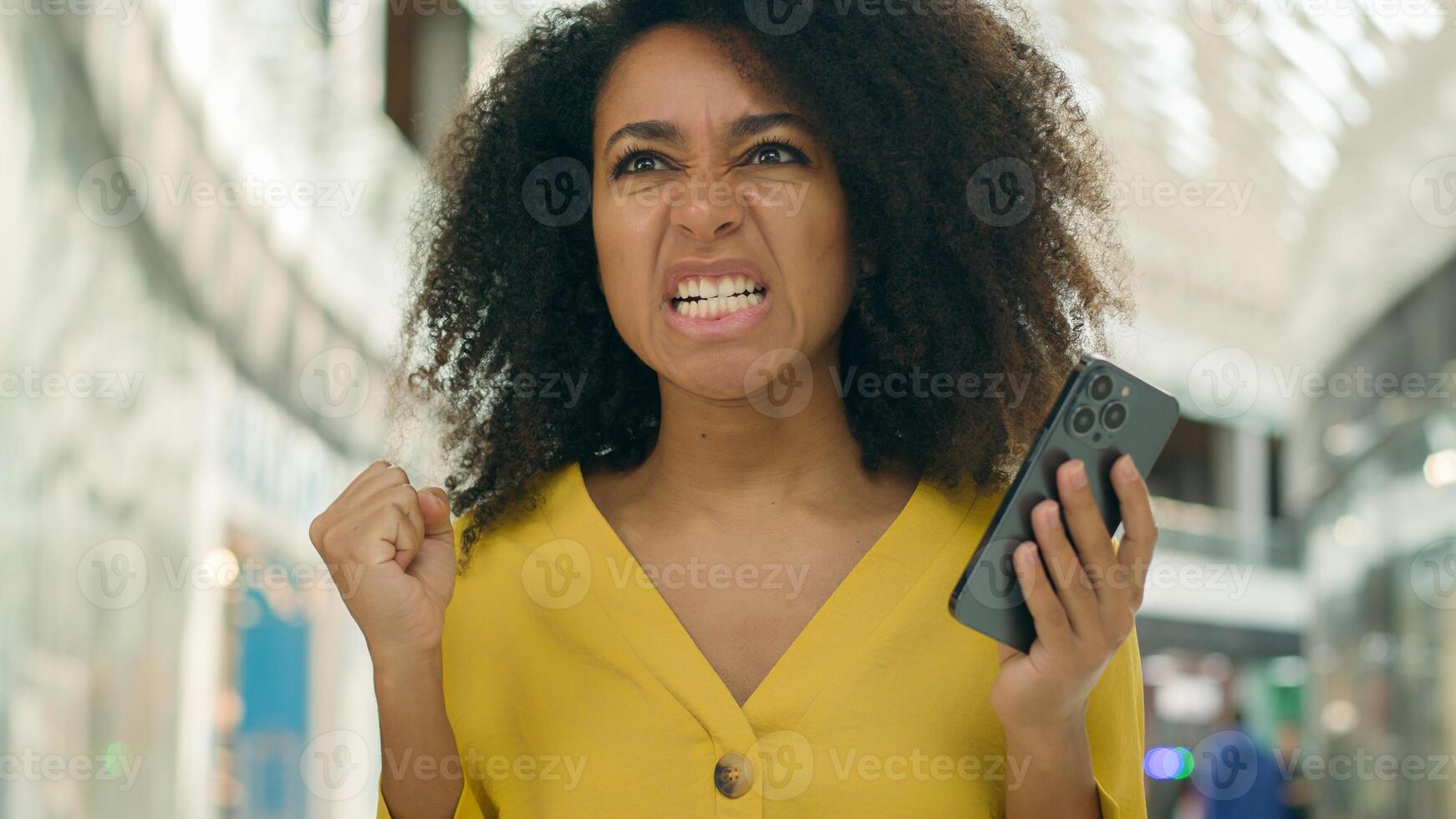 Young unhappy African American woman looking at screen display smartphone telephone error failure evil angry female screaming problem trouble breaking dissatisfied bad news messages annoyed technology photo