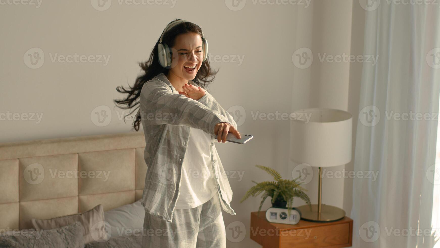 Cheerful woman jumping dancing on bed bedroom home mobile phone enjoy joyful headphones listening music female technology singing happy excited energy pastime celebrate active leisure moving apartment photo