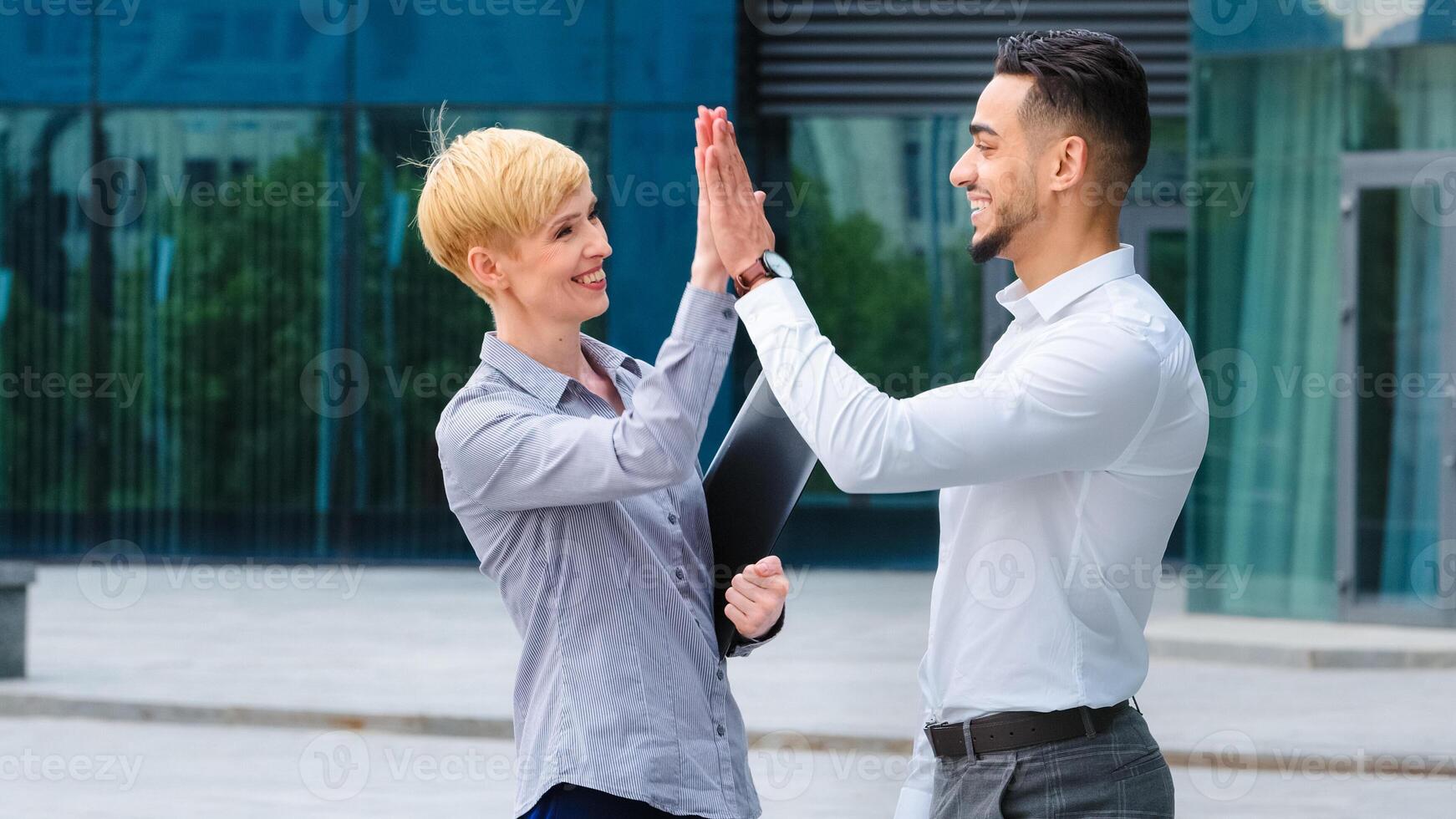 Multi-ethnic co-workers Caucasian woman and Hispanic man stand outdoors company building make high five. Businesswoman with folder discussing project with businessman agree teamwork gesture success photo