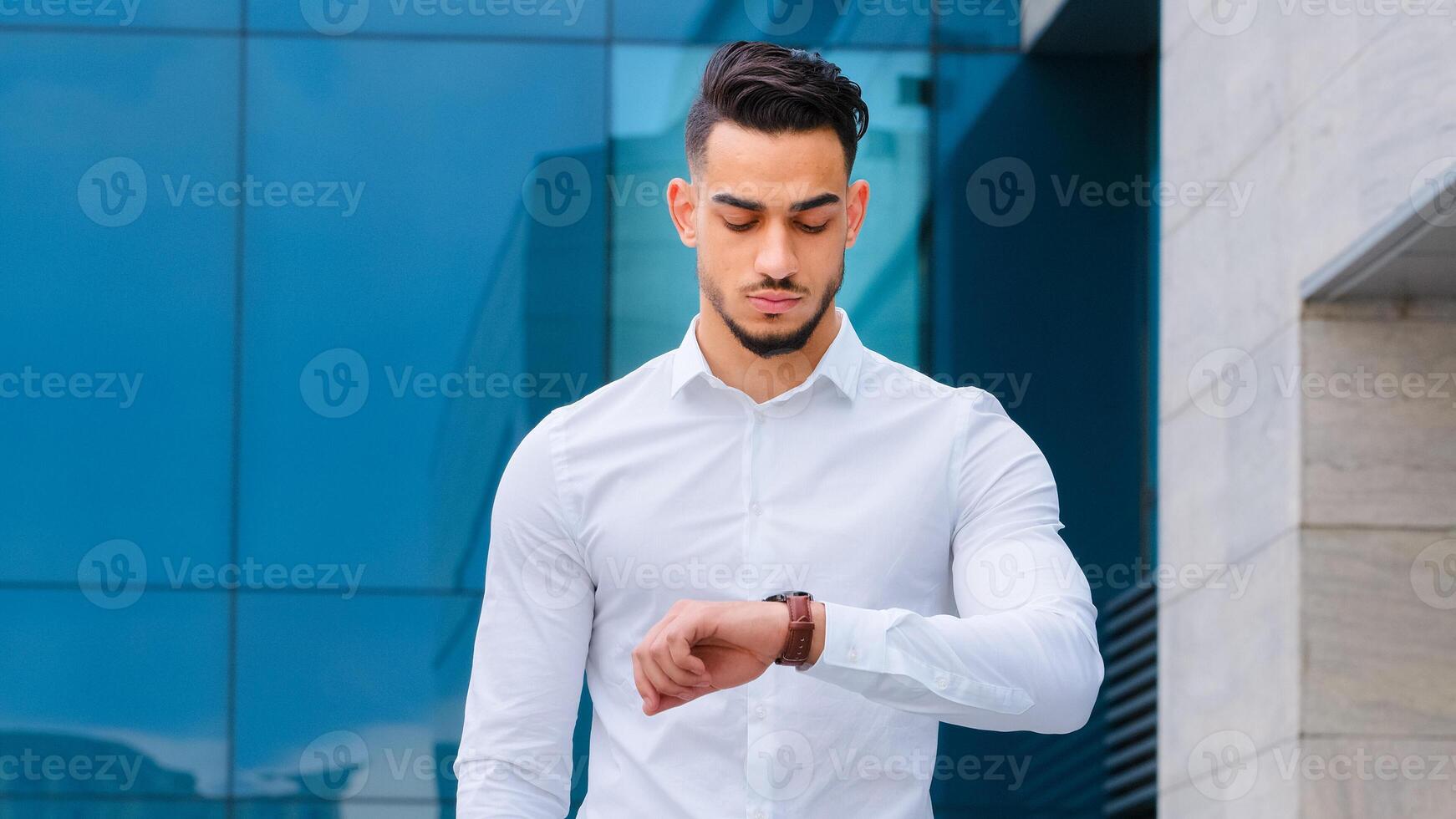 Serious upset angry hispanic indian man bearded guy disgruntled businessman boss wears formal stylish shirt stands outdoors looking at wrist watch worried about being late lateness time rush waiting photo