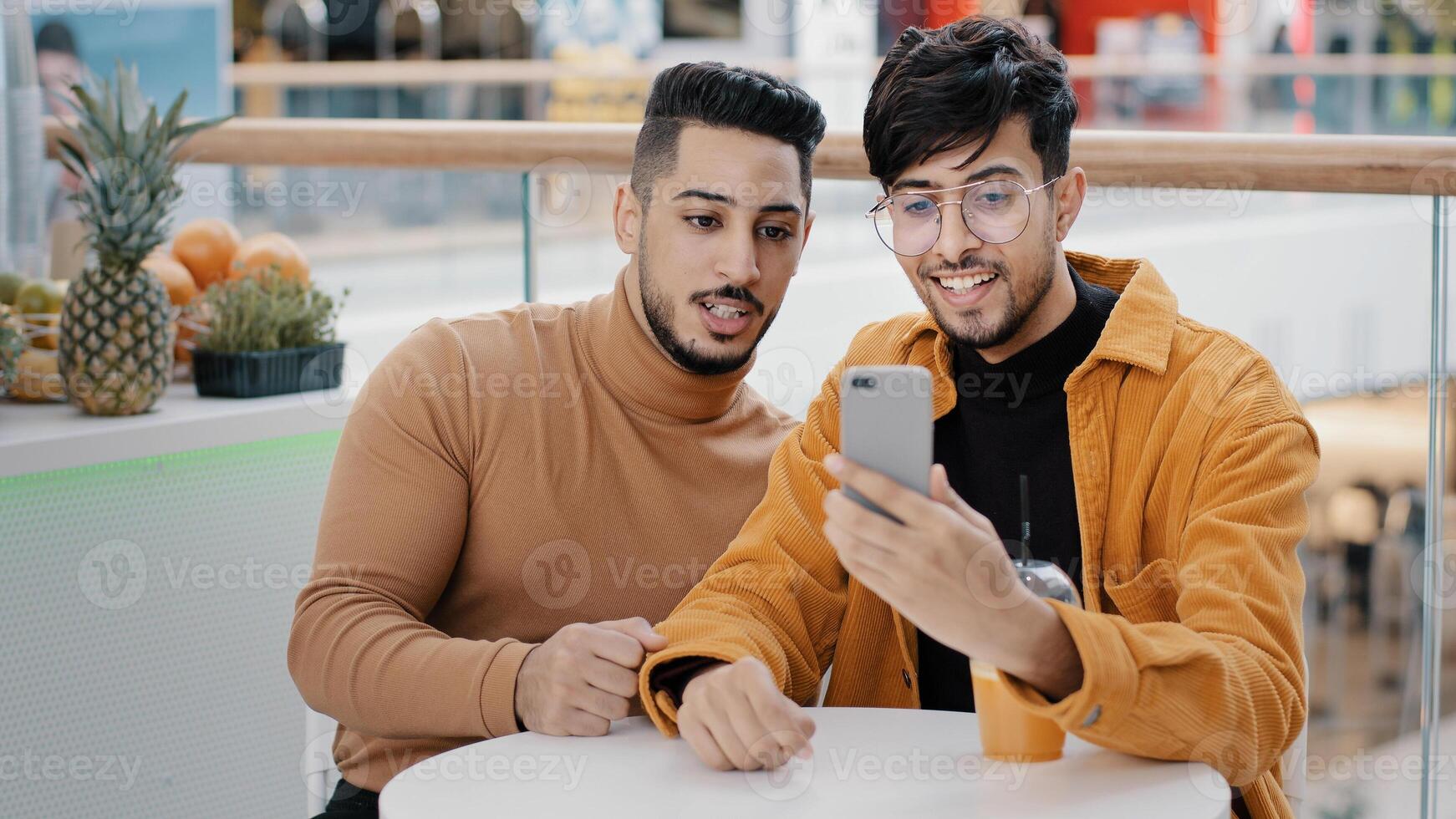 Two happy arab friend bloggers sitting in cafe recording blog on phone communicating via webcam looking at smartphone screen take selfie making videos on social media using mobile application photo