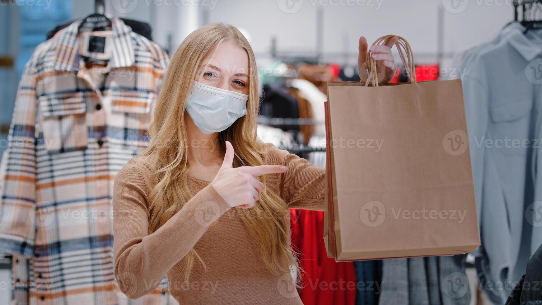 Close-up happy Caucasian woman in medical mask covid pandemic shopping stand in clothing store shop girl consumer showing pointing on bags purchases lady satisfied low prices discount Black Friday photo