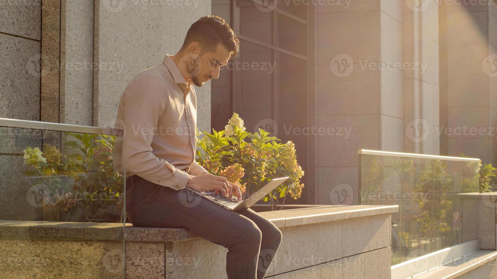 Hispanic businessman sitting outside with laptop working remote distant job outdoors near office company building. Young Arabian user student worker typing computer app in city online chatting in net photo