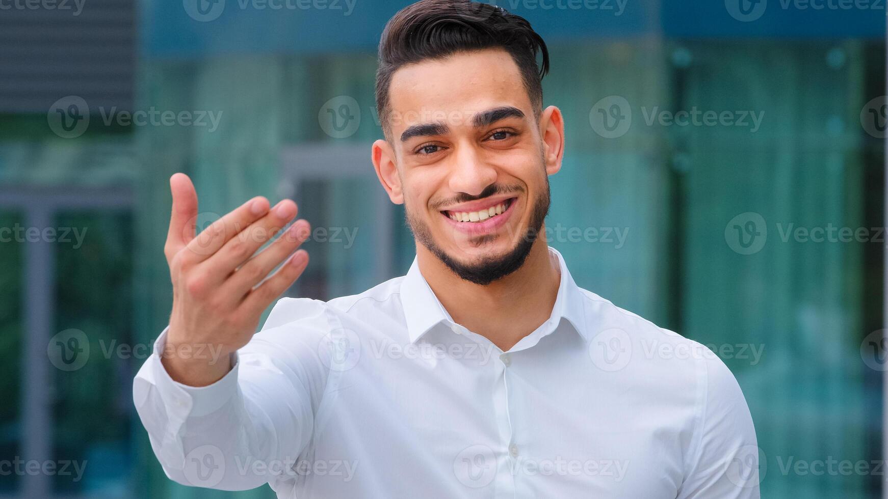 Portrait friendly smiling toothy hispanic arab business man spaniard boss wears white formal shirt showing gesture of invitation advice to approach with hand asks to come outdoors, body languages sign photo