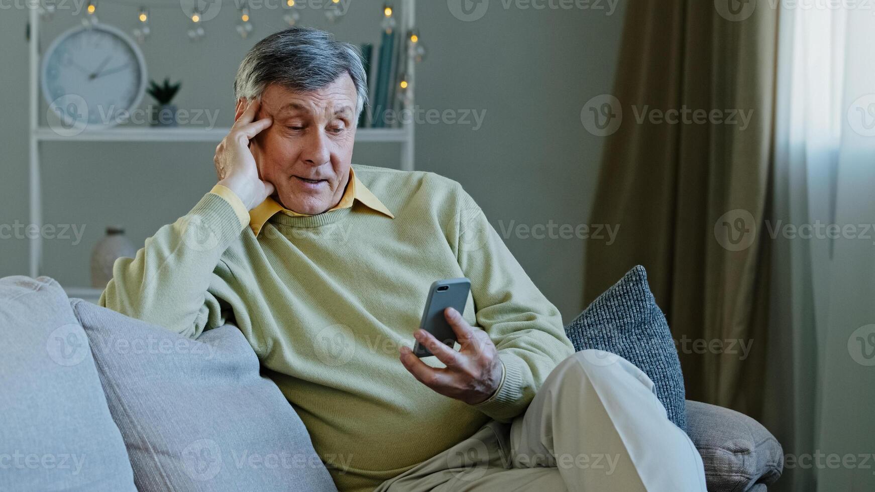 Happy elderly adult man sitting on sofa hold gadget studying app on smartphone makes online order smiling grandfather resting in room reading news using phone old people with modern technology concept photo