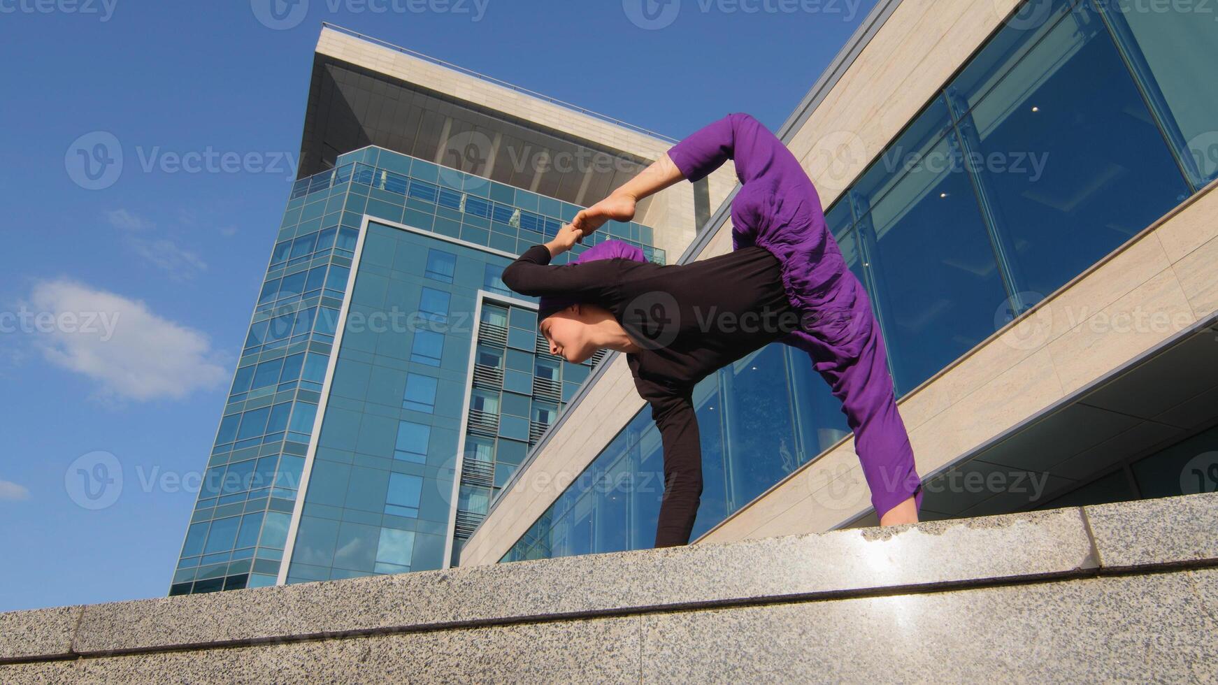 Muslim islamic girl woman yogi sports athlete stands urban background city building doing stretching workout yoga holds leg in air holding foot high with hand balance asana pose concentration concept photo