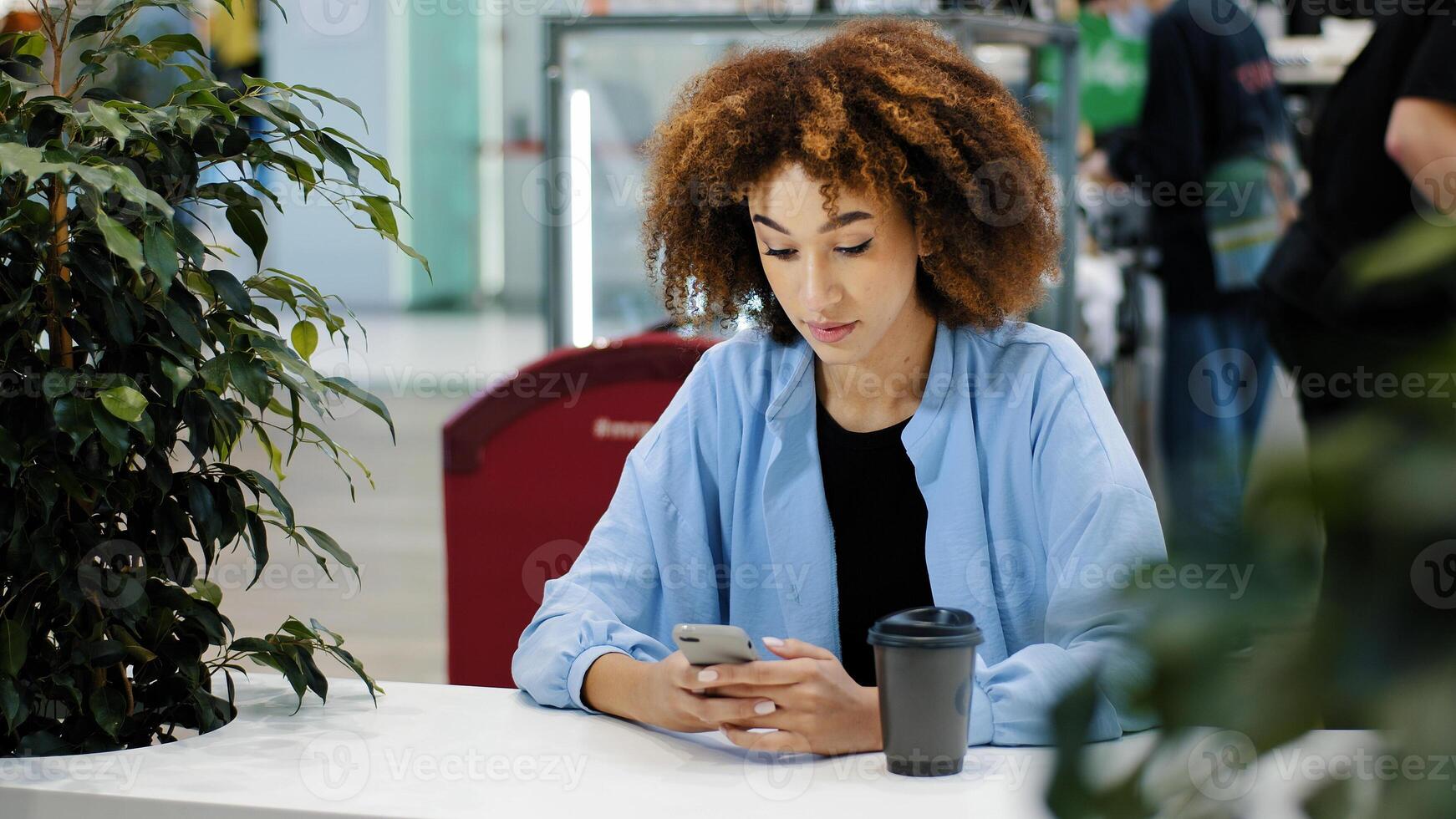 African 20s woman multiracial biracial lady with curly haircut sitting at cafe table drinking coffee tea from paper cup looking in mobile phone typing smartphone chatting browsing buying online app photo