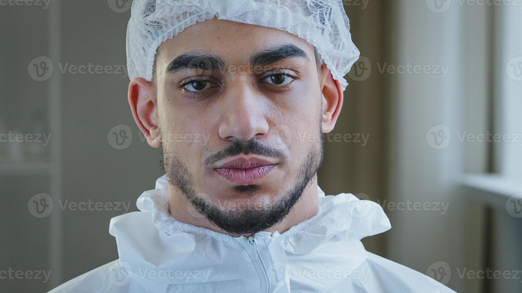 Portrait of exhausted tired young arab male doctor nurse practitioner in protective uniform latex gloves stands in clinic takes off medical cap from head exhales with relief after hard day at work photo