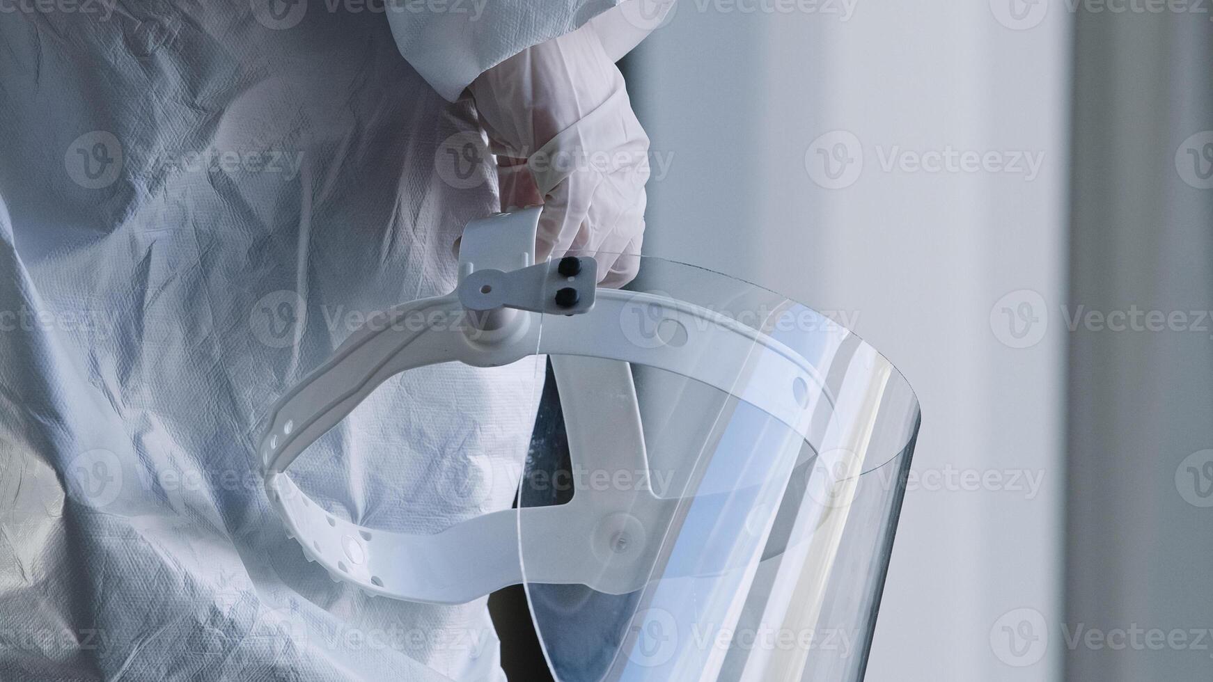 Unrecognizable doctor medical worker nurse practitioner scientist stands in clinic laboratory hospital at window wears white uniform and latex gloves holds in hand transparent protective face shield photo