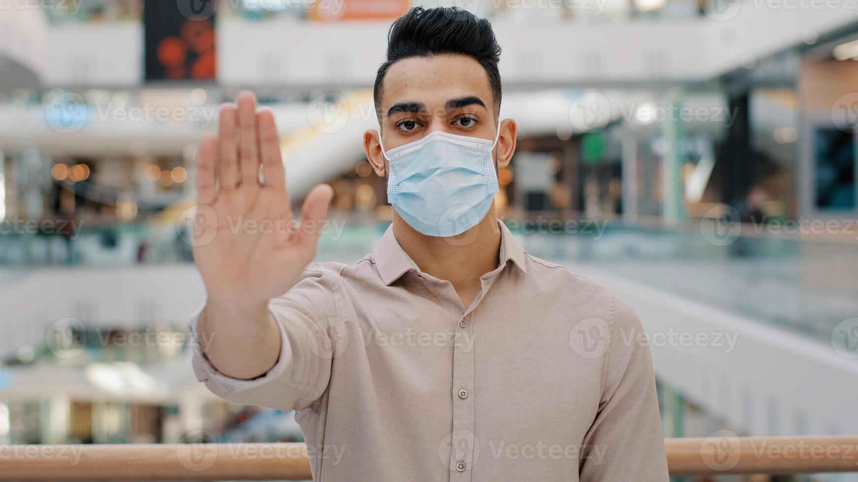 Portrait of young hispanic man wearing medical mask put palm forward serious ethnic guy looking at camera male showing stop sign palm with hand gesture safe distance coronavirus epidemic protection photo