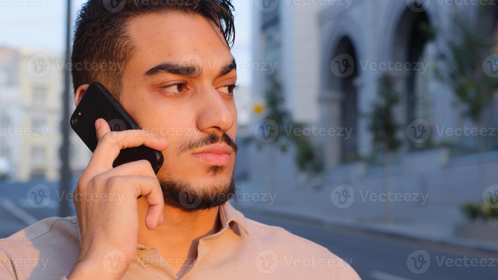 Close-up hispanic emotional business man bearded serious guy talking on mobile phone answering call using smartphone remote talk cellular connection standing in city outside cellphone negotiation photo