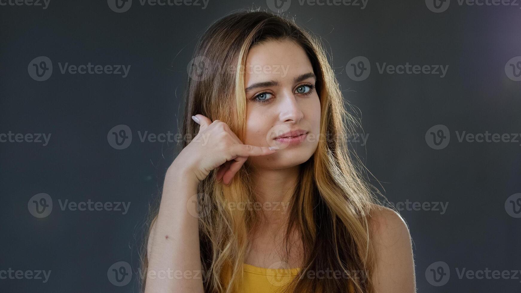 Young lady girl invites contact by phone. Playful happy pretty woman making telephone gesture near head call me sign calling symbol offering communication posing in studio isolated on gray background photo