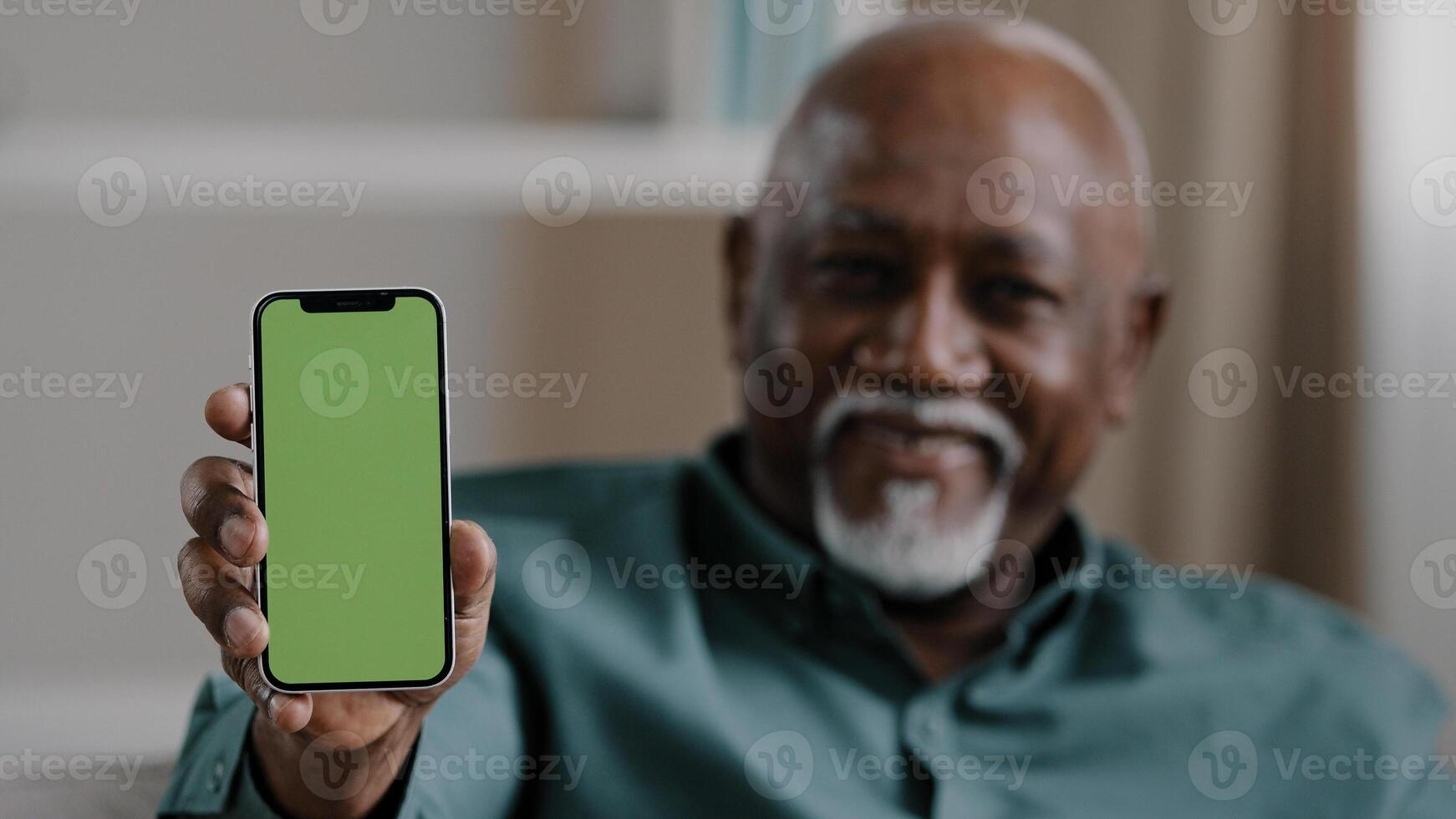 Old male African American biracial man at home sitting on couch businessman in office showing smartphone with empty green screen chromakey holding phone shows to camera advertisement gadget touchpad photo
