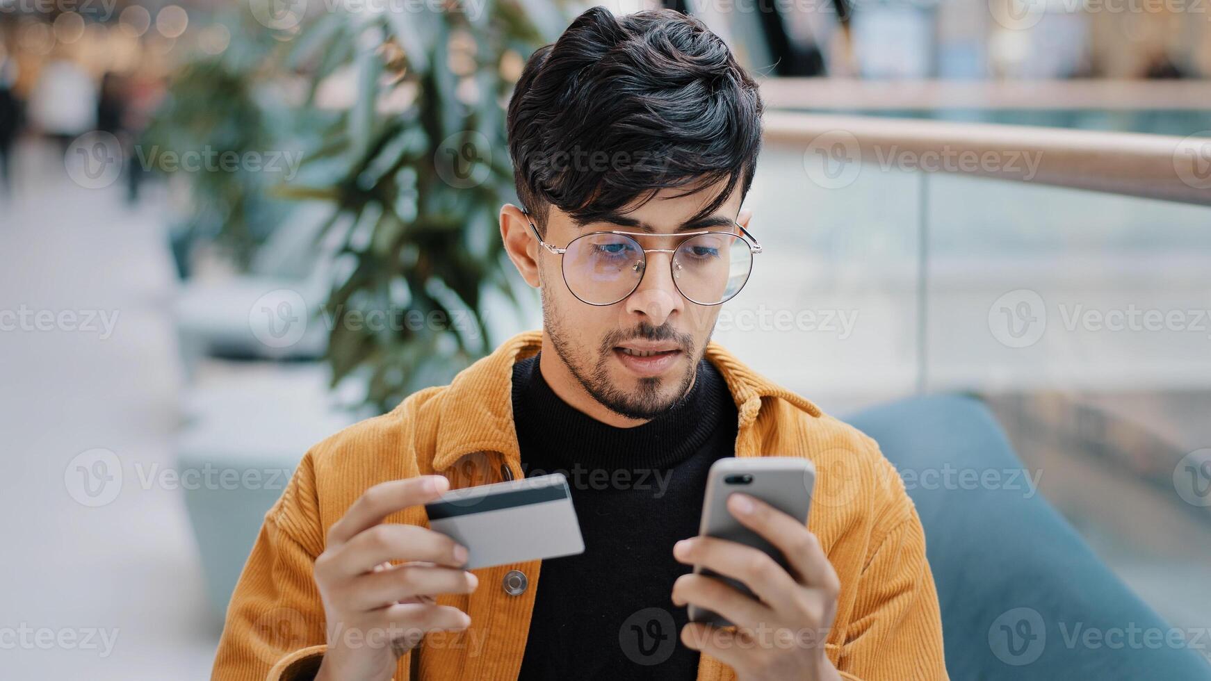 Focused indian man guy customer pay by card on phone using e-banking mobile application arabian male shopper making buying in online store paying purchase use instant easy payment on smartphone photo