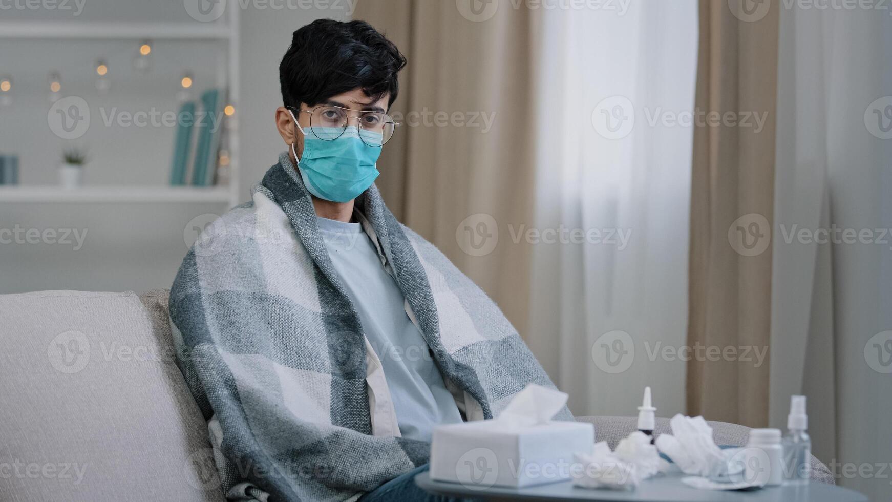 Sick arab man in medical face protective mask sitting on couch at home ill guy wrapped in plaid looking at camera with sadness suffering seasonal virus covid coronavirus pandemic contagious infection photo