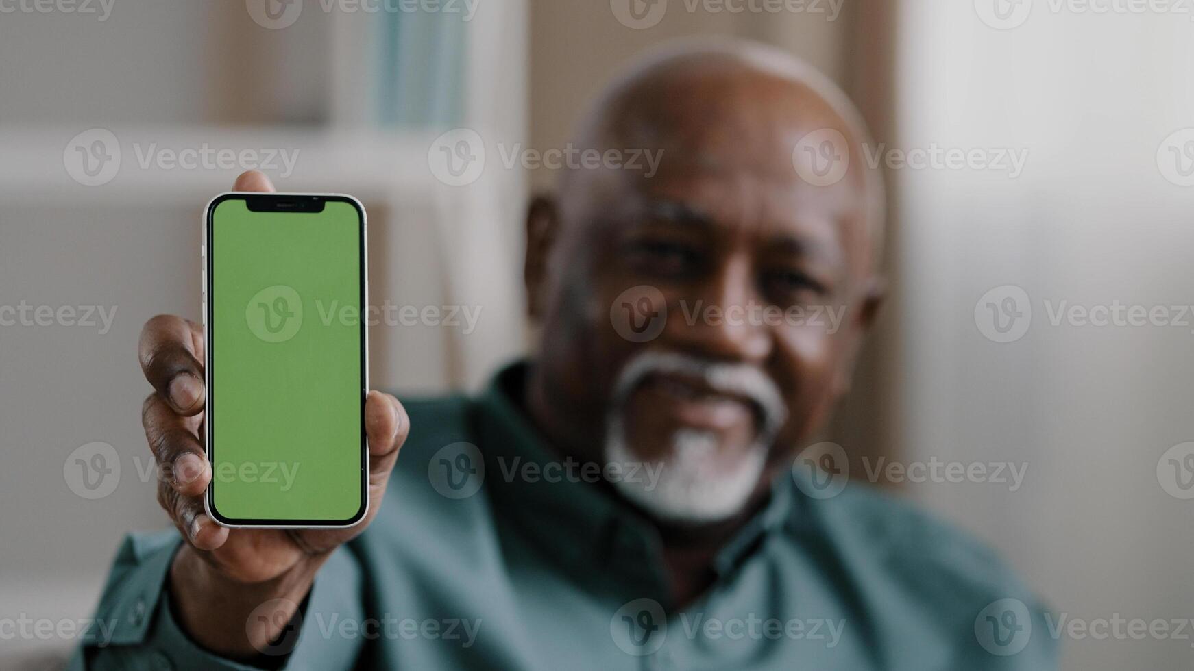 Old male African American biracial man at home sitting on couch businessman in office showing smartphone with empty green screen chromakey holding phone shows to camera advertisement gadget touchpad photo
