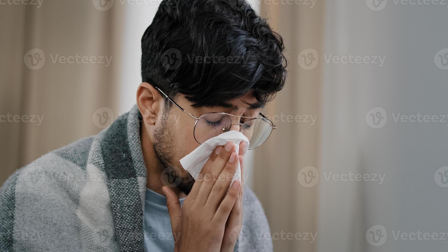 Close-up sad exhausted arabic bearded man suffering from seasonal allergy dust virus respiratory disease runny nose guy in glasses wrapped in blanket sitting at home blowing nose snot in paper napkin photo