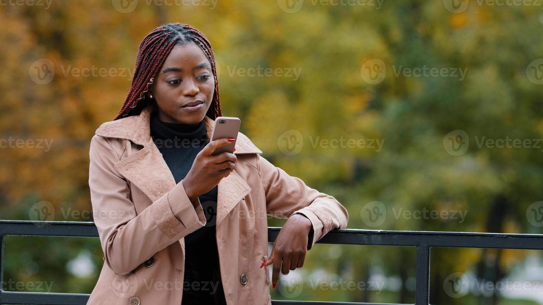 Young serious female standing in autumn park holding mobile phone browsing email remotely communicating in social network using smartphone outdoors searching for information on internet writes sms photo