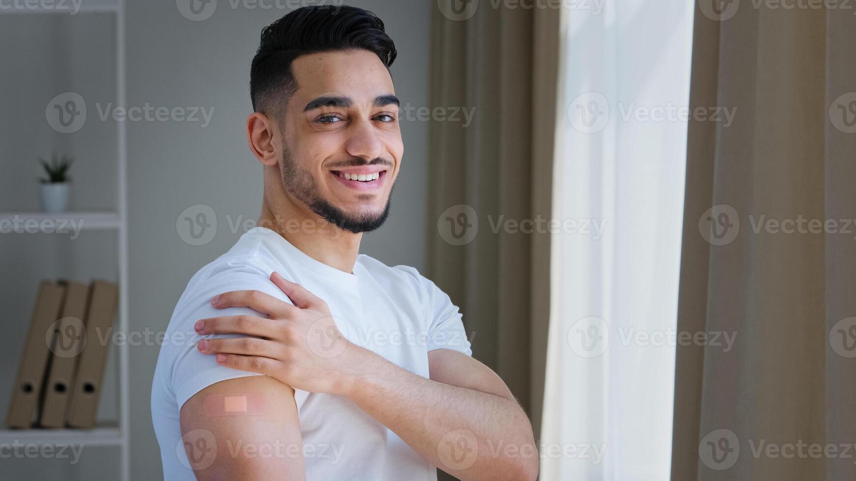 Happy satisfied indian guy arab man hispanic patient male posing indoors wears white t-shirt shows plaster on shoulder feels relief after vaccination injection with covid remedy medical immunization photo