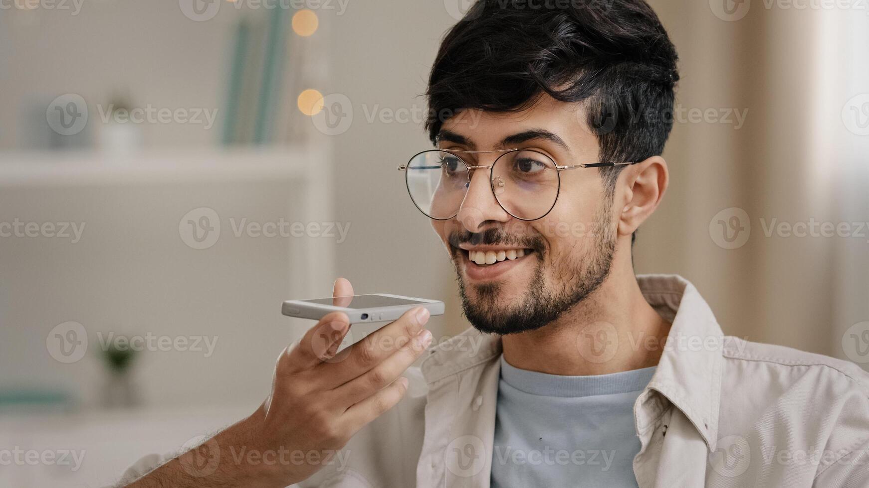 Friendly young arab man recording voice recognition message on speakerphone sitting at home. Bearded guy with glasses using virtual assistant app sets reminder on mobile phone indoors dictates words photo