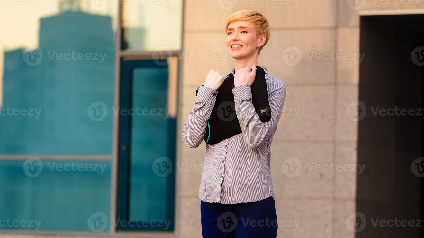Happy mature adult business woman boss leader stands outdoors near company office building holds laptop in case hugs embraces computer rejoices at start inspired by of work after lockdown quarantine photo