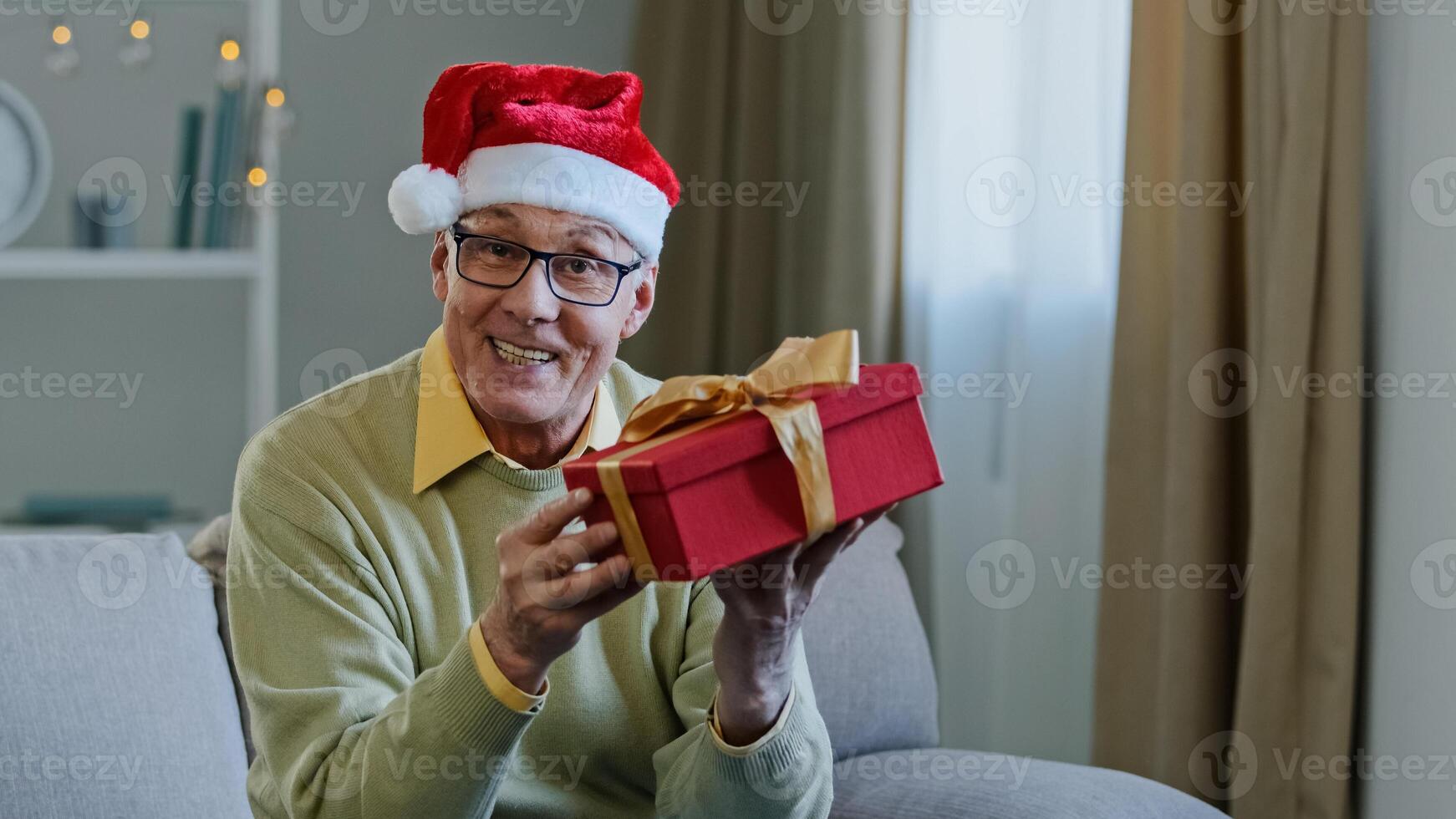 Old Caucasian senior mature man grandfather in Santa Christmas hat at home singing song dancing moving hands having fun celebrating x-mas New Year party showing gift box present talking call photo