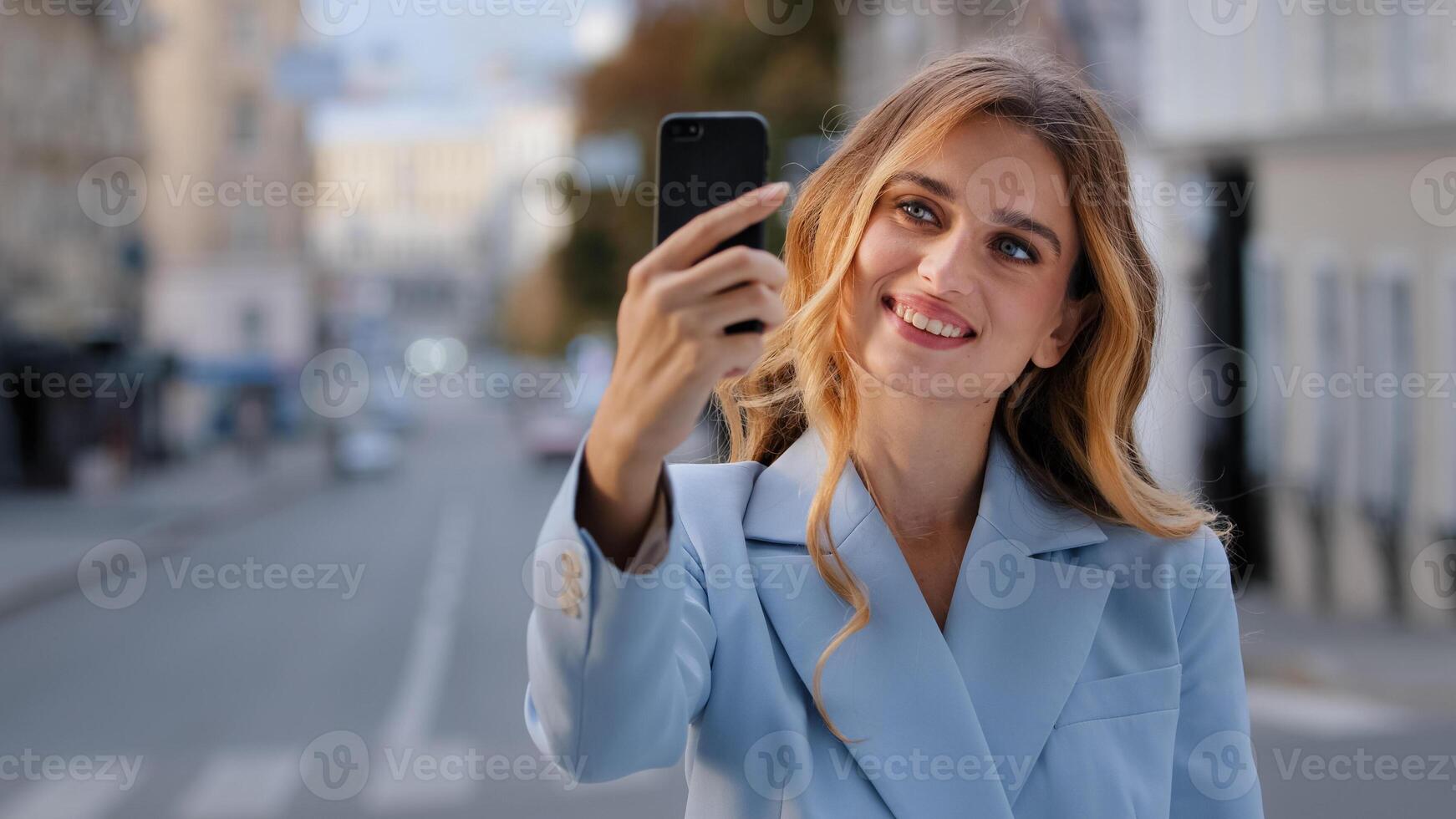 Portrait of young beautiful girl Caucasian business woman blogger vlogger model stands in city holds phone taking photo on mobile camera shoots for vlog blog with smartphone online chat call