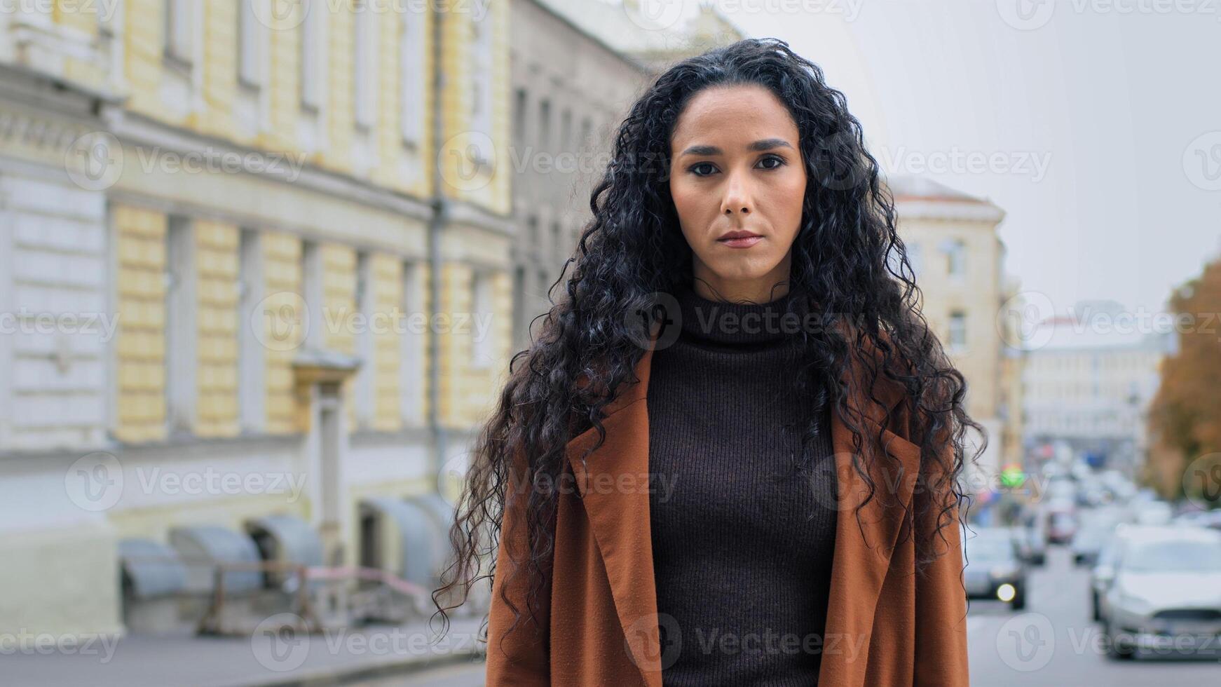 Serious alone confident powerful young brunette girl with long curly hair business woman with angry expression stands on street in city looking at camera intently gaze, loneliness problem concept photo