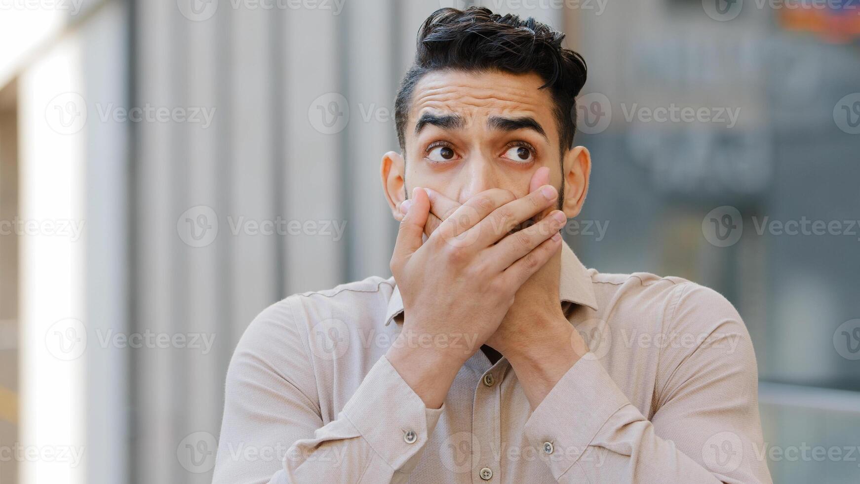 Confused afraid scared young guilty man shocked surprised guy male hispanic arab indian covers mouth with hands worried about telling secret feels shock bad news looking at camera, problem concept photo