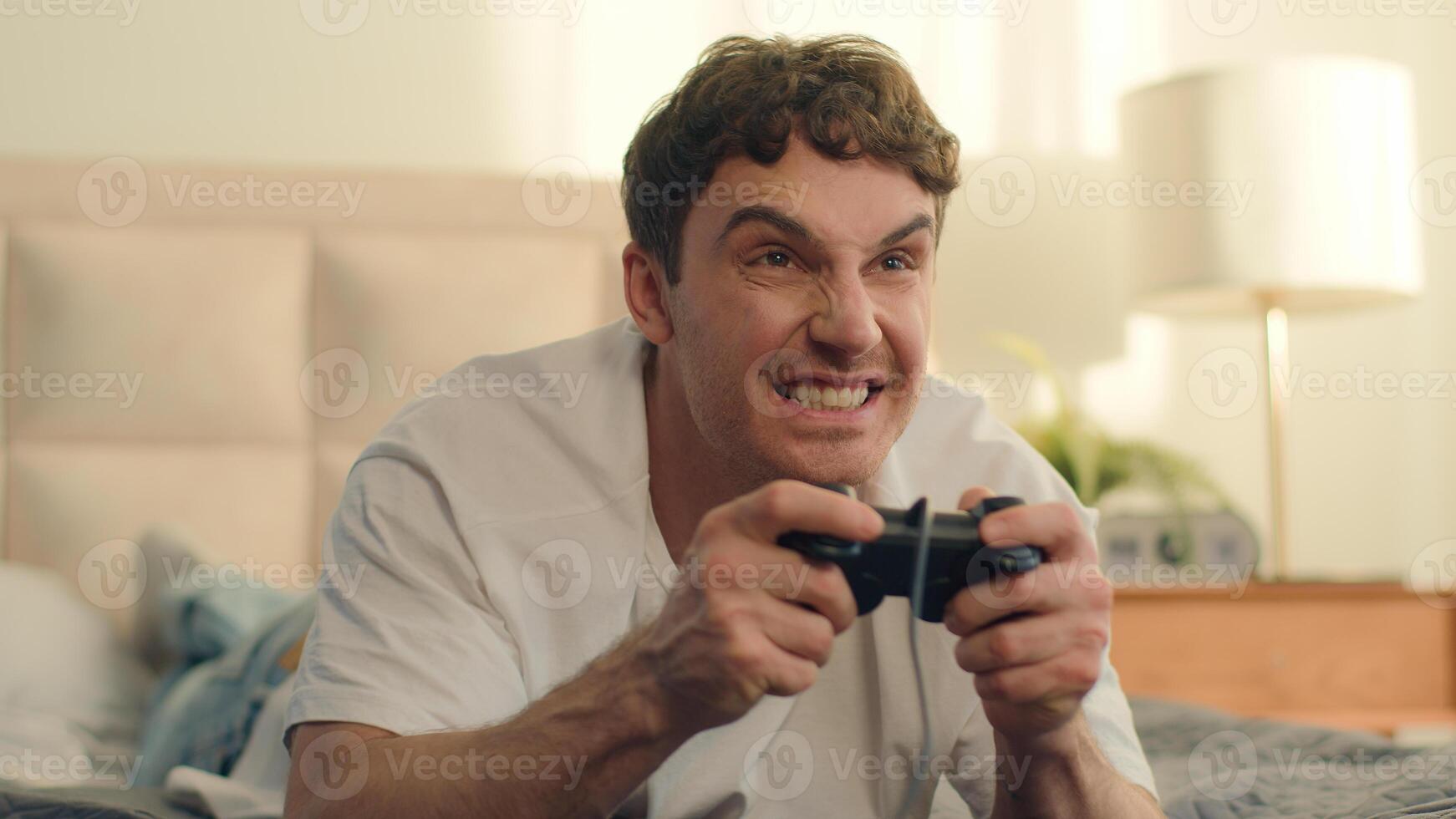 Playful Caucasian European young man lying on bed playing console holding joystick games fun weekend home enjoy guy hobby leisure hands technology virtual cyberspace cheerful excitement smiling photo