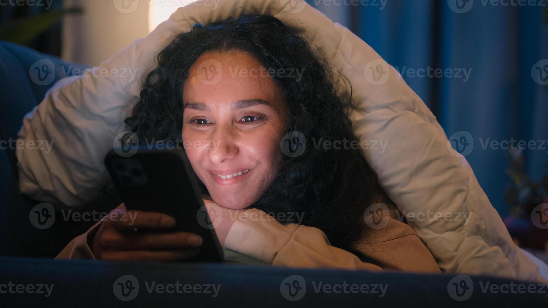 Happy woman gadget addict social media addicted Hispanic smiling girl Latino Indian lady Arabian female suffering insomnia at night home relaxing under blanket cover with duvet browsing mobile phone photo
