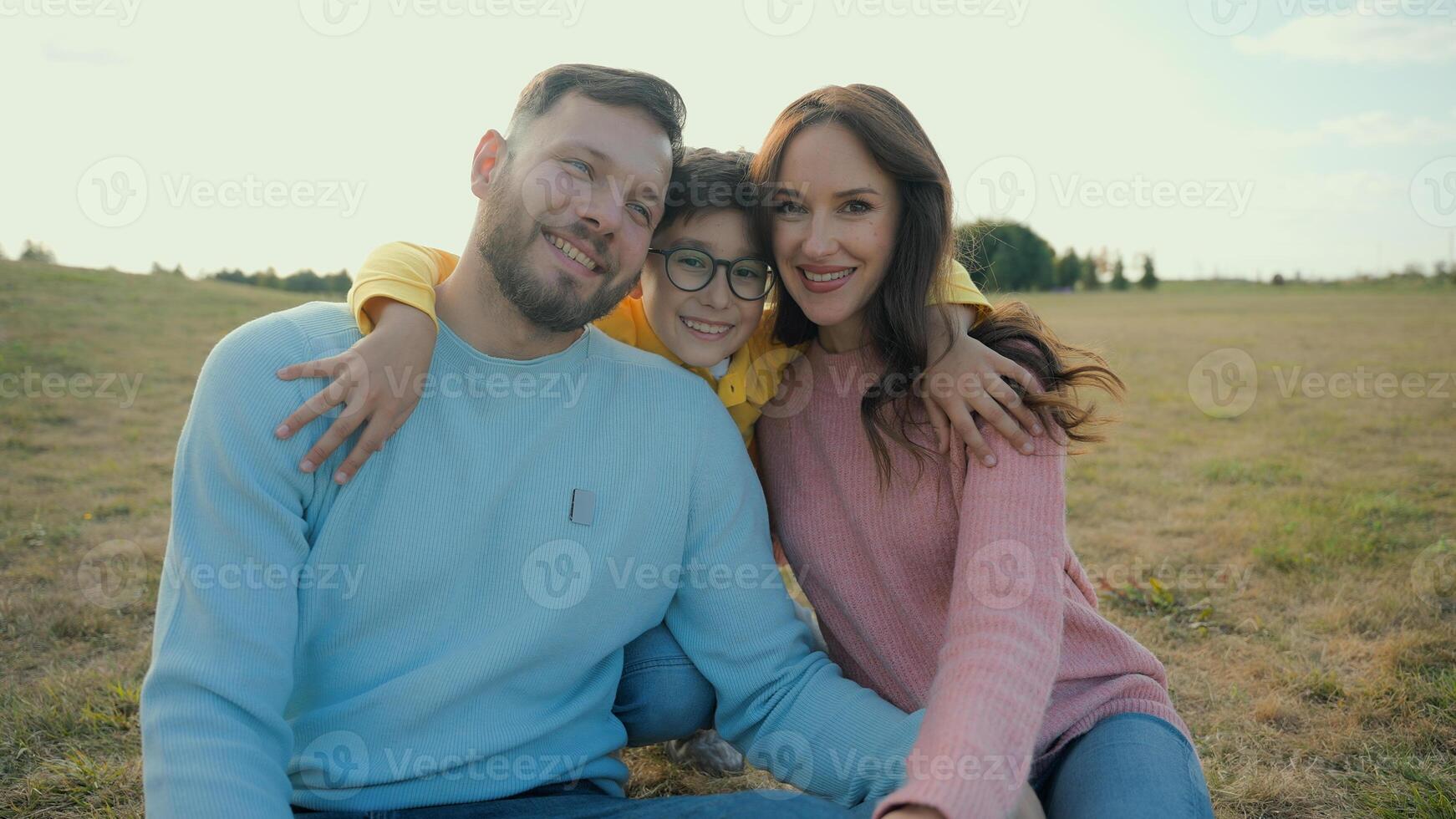 Joyful Caucasian family picnic married couple mother female woman father dad guy man male hug happy little boy child kid son walking to parents embrace cuddle together looking at camera nature meadow photo