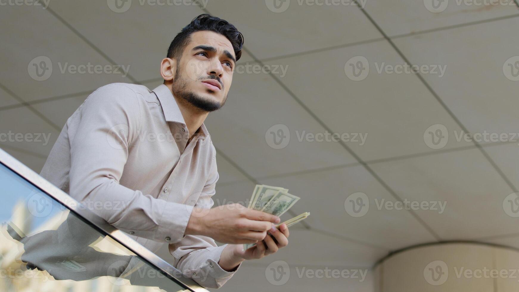 Bottom view young pensive successful bearded guy rich business man holding bundle of money banknotes dollars money thinking about finances credit profit salary loan investments standing on balcony photo