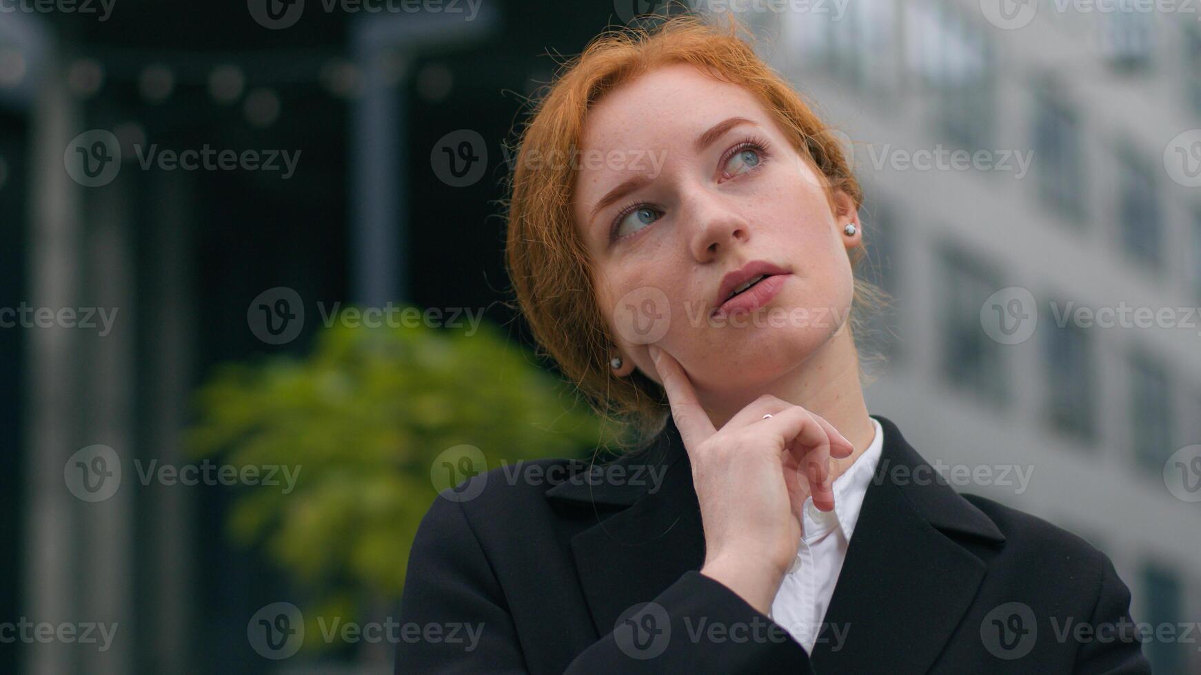 Close up young puzzled Caucasian ginger businesswoman girl female woman student elegant suit think finger touch cheek contemplating gesture outdoors city street. Business choice decision idea planning photo