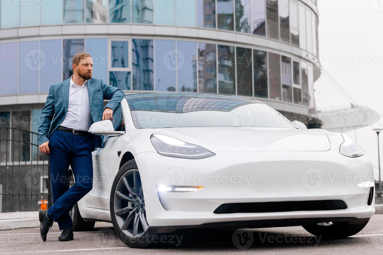 A middle-aged businessman charges an electric car from a charging station powered by renewable energy sources against the backdrop of the business center photo