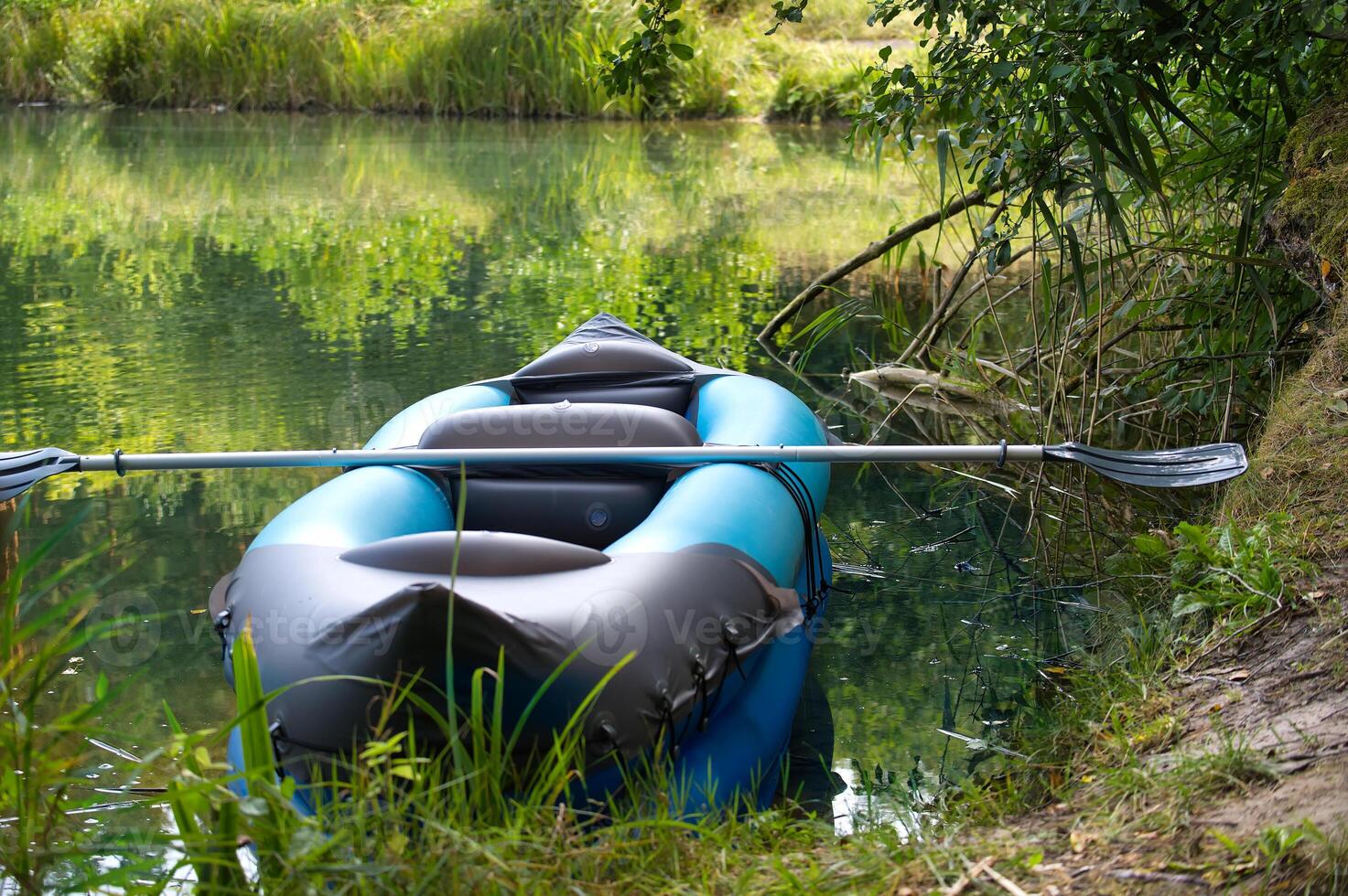 Inflatable kayak with paddle resting on serene lakeshore surrounded by lush green trees photo